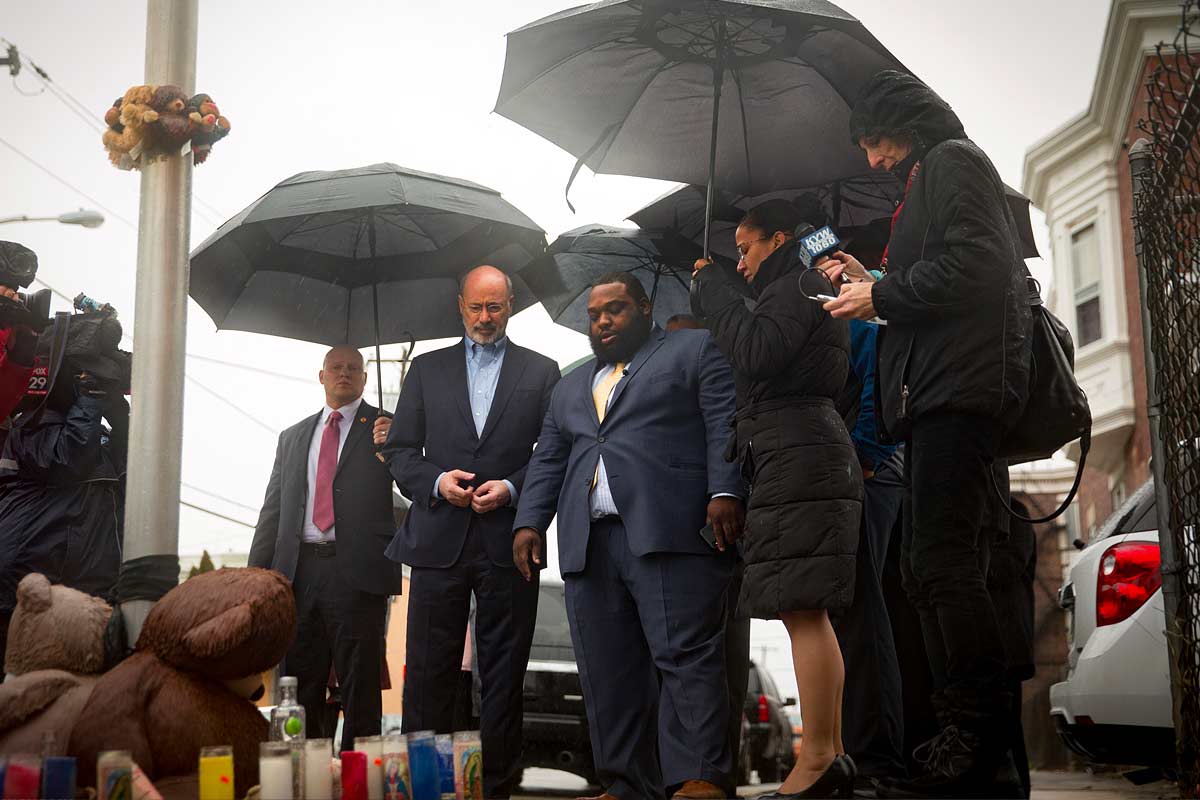 Elected officials standing in the rain looking a street memorial for a child killed by gunfire